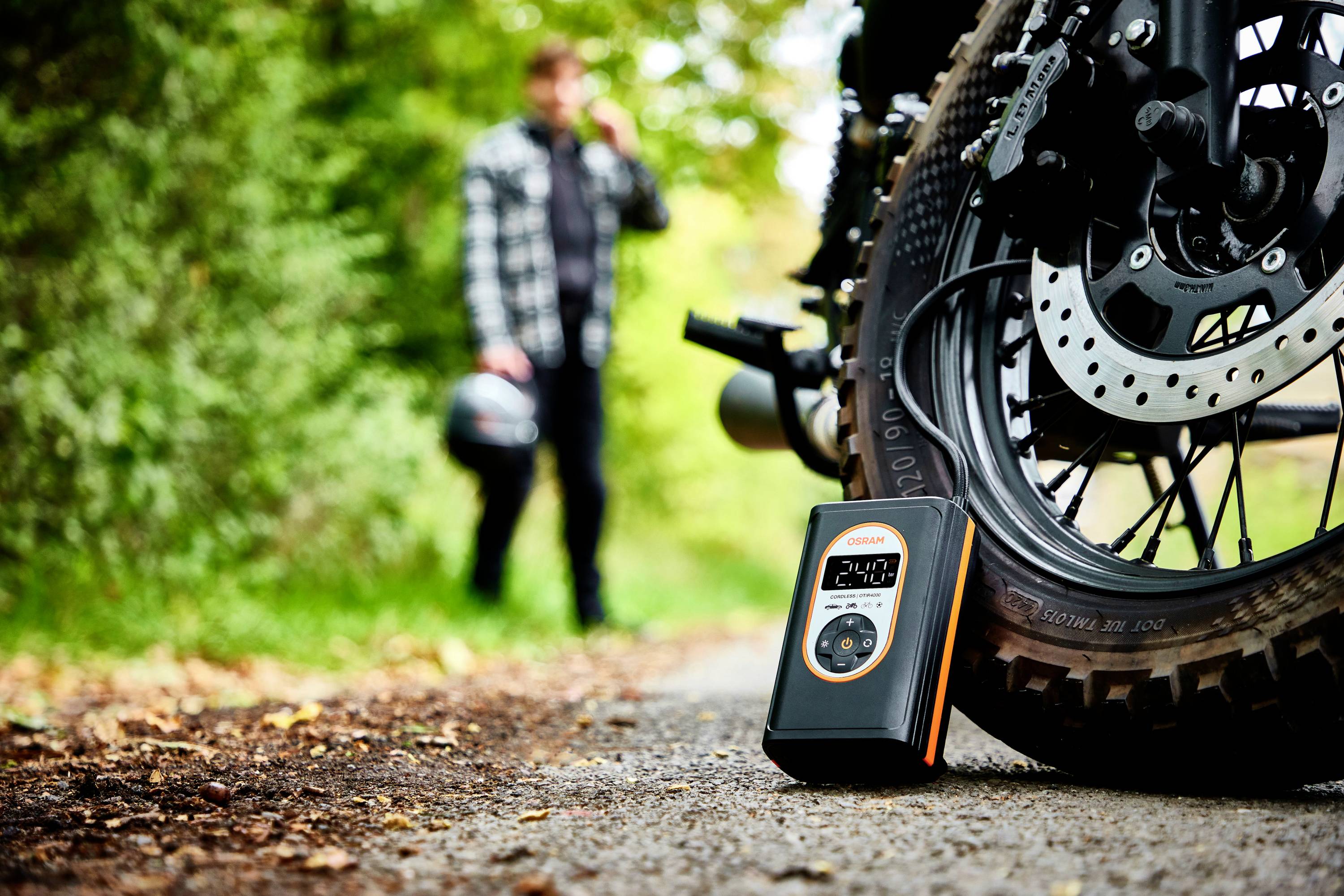 Un manomètre de pression des pneus est posé à côté d'une moto sur un chemin forestier. En arrière-plan, une personne floue se tient avec un casque à la main.