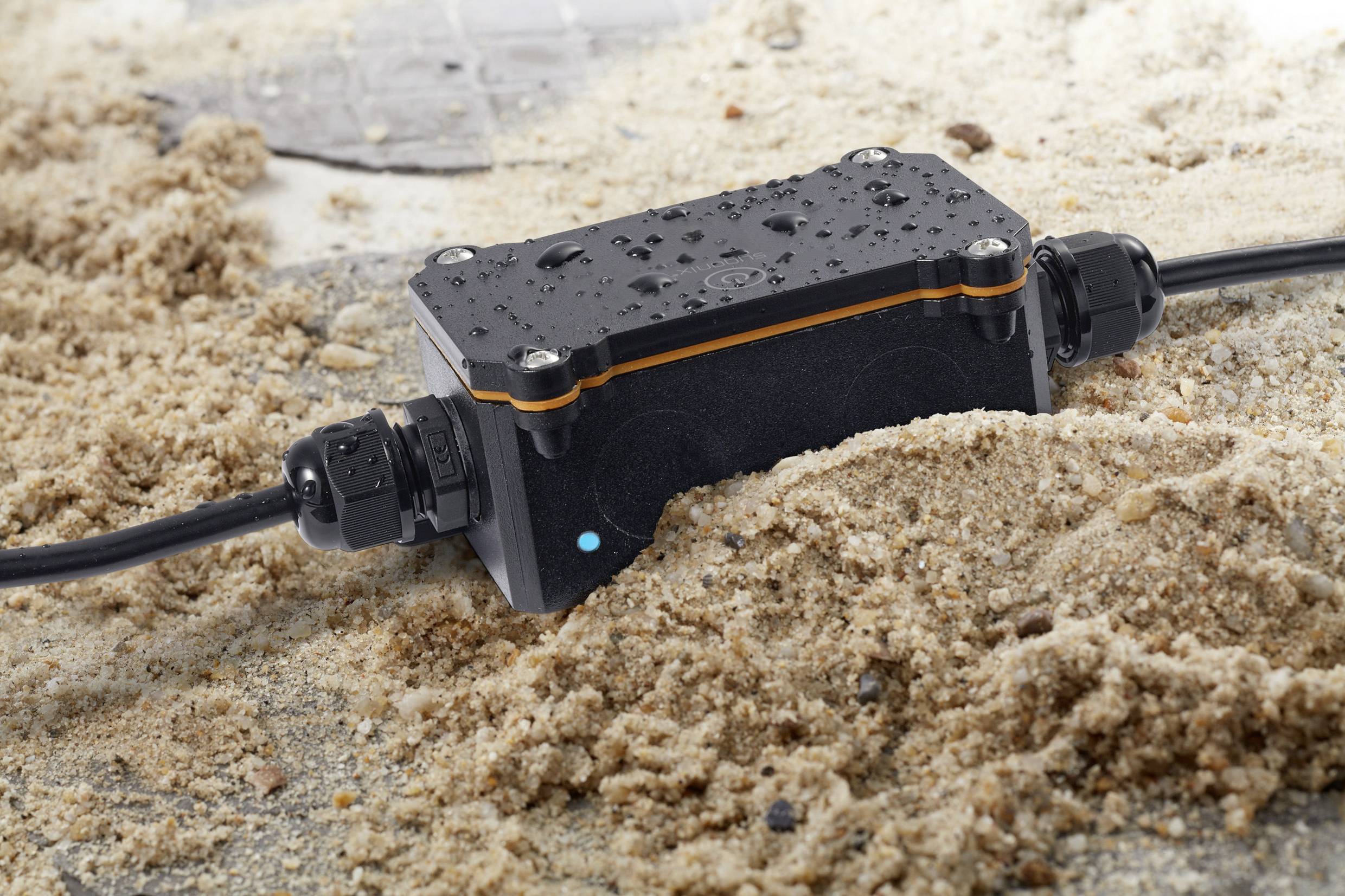Un boîtier de raccordement électrique noir et étanche repose dans le sable, avec des gouttelettes d'eau et des câbles connectés à ses extrémités.