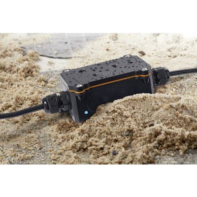 Un boîtier de raccordement électrique noir et étanche repose dans le sable, avec des gouttelettes d'eau et des câbles connectés à ses extrémités.