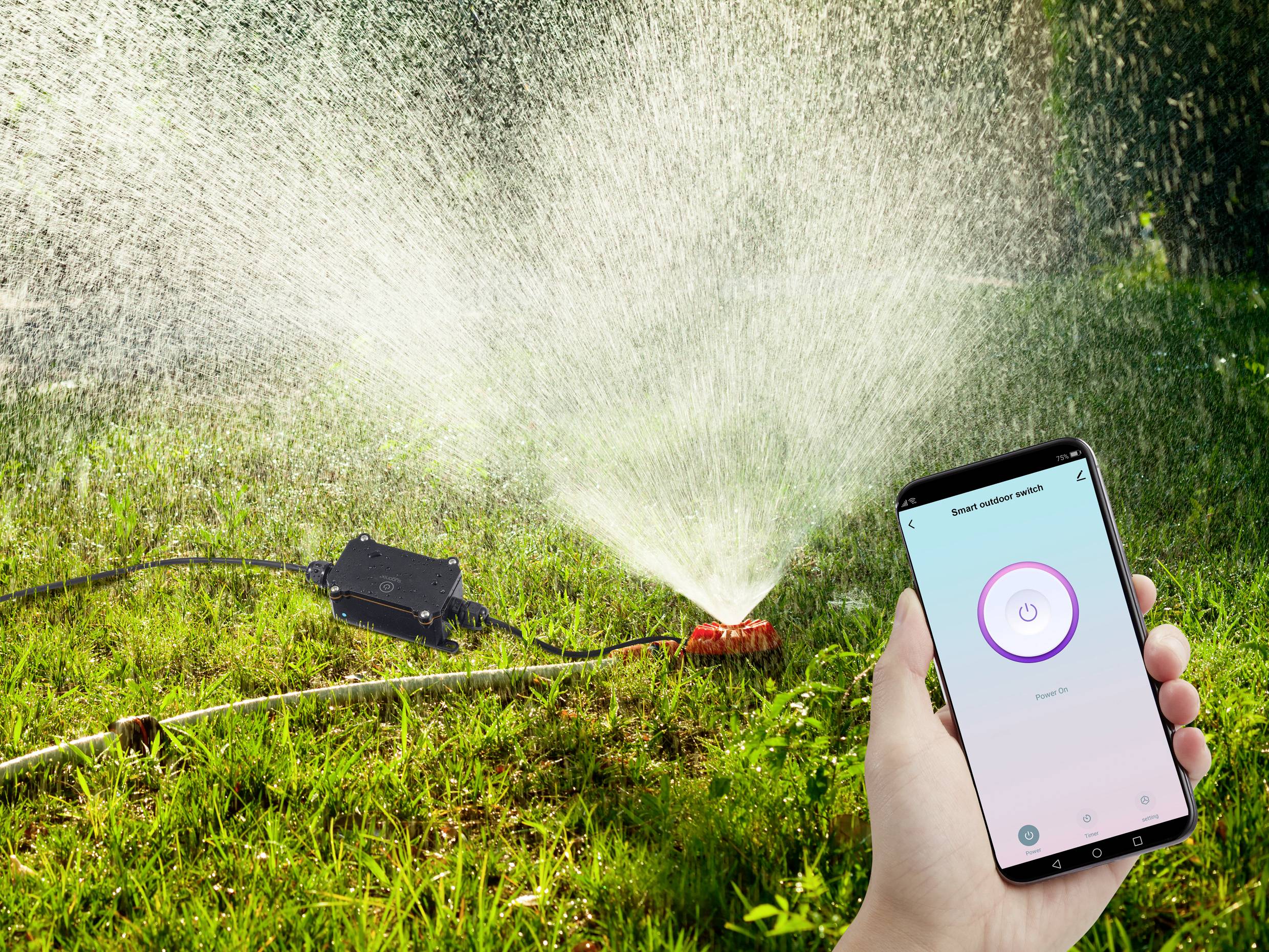 Une main tient un smartphone affichant une application de commande d'un arroseur automatique. L'arroseur automatique irrigue la pelouse.
