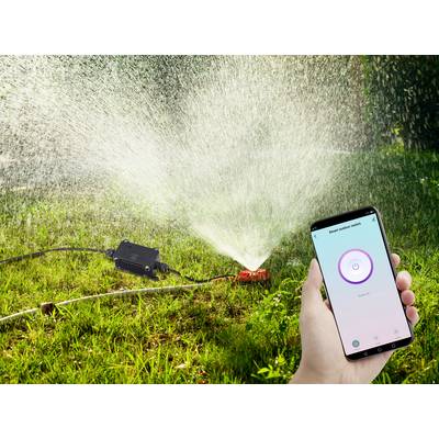Une main tient un smartphone affichant une application de commande d'un arroseur automatique. L'arroseur automatique irrigue la pelouse.
