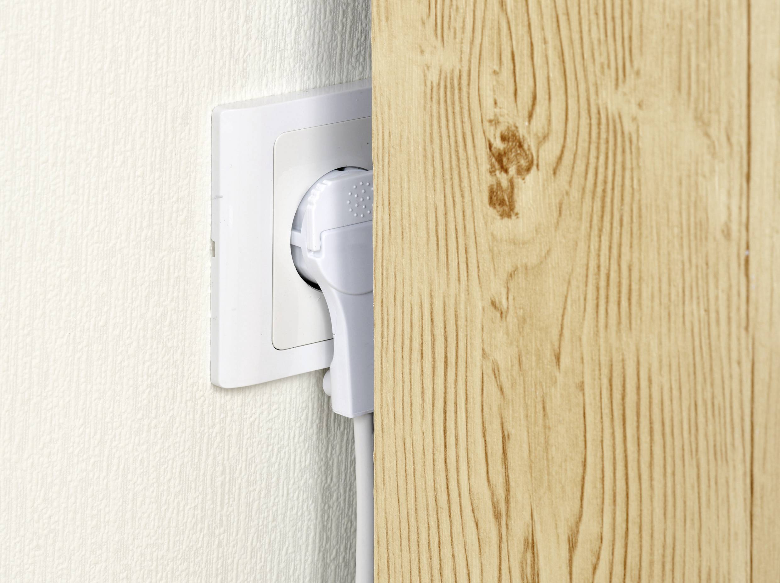 Une fiche blanche est branchée dans une prise murale, avec une planche de bois qui bloque partiellement l'accès.