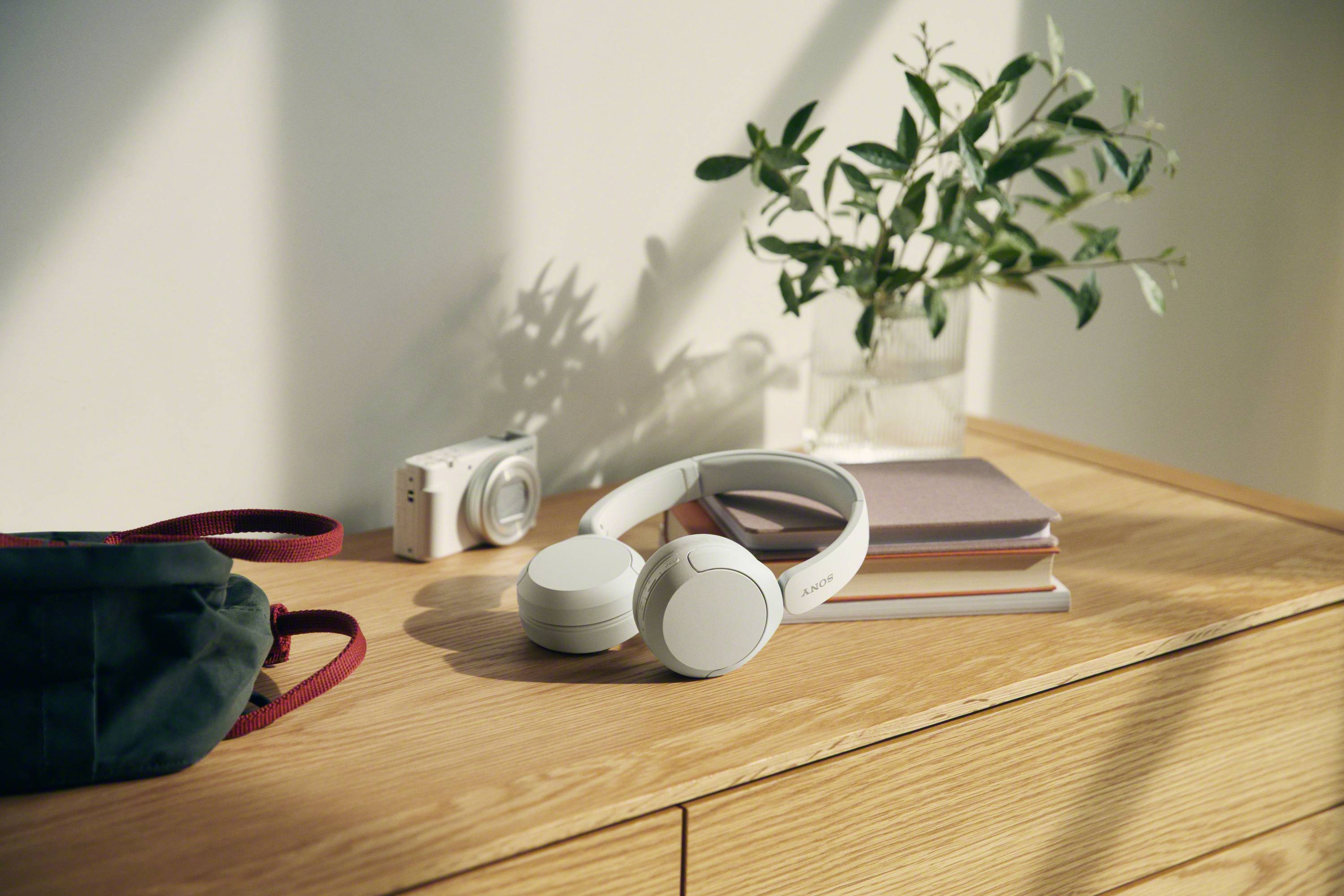 Casque blanc posé sur une pile de livres sur une table en bois, à côté d'un appareil photo et d'une plante ; atmosphère détendue.