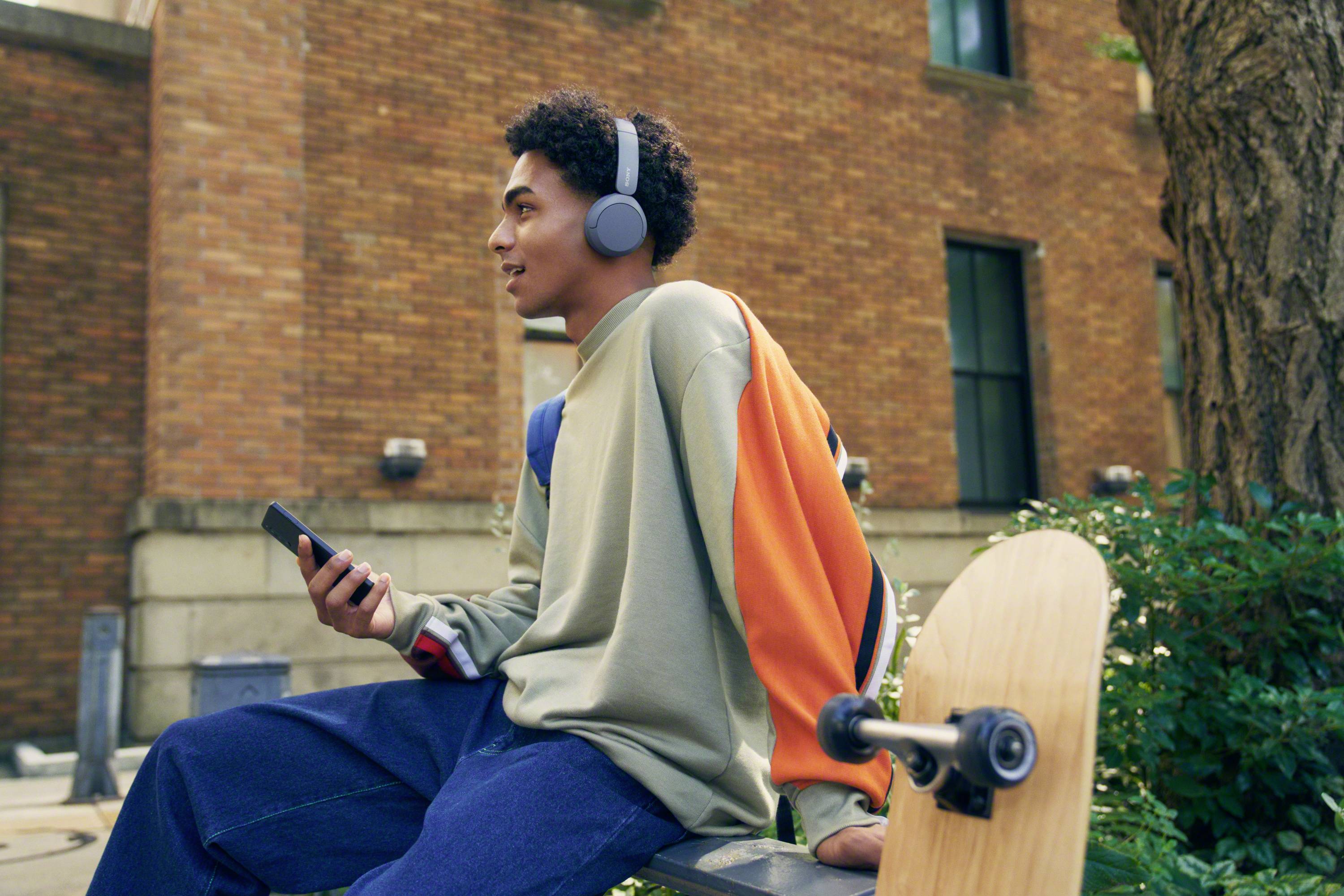 Une personne portant des écouteurs est assise sur un banc devant un bâtiment, avec un smartphone et une planche de skateboard.