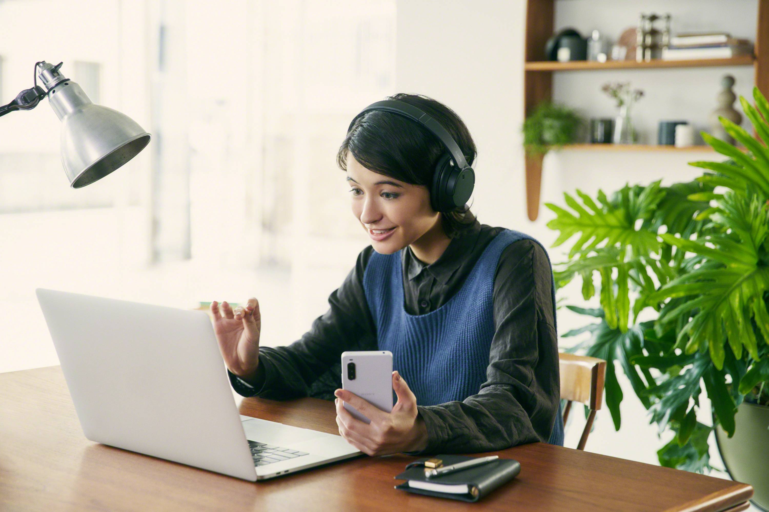 Une personne souriante portant des écouteurs est assise à une table, utilisant un ordinateur portable et tenant un smartphone. Des plantes sont visibles en arrière-plan.