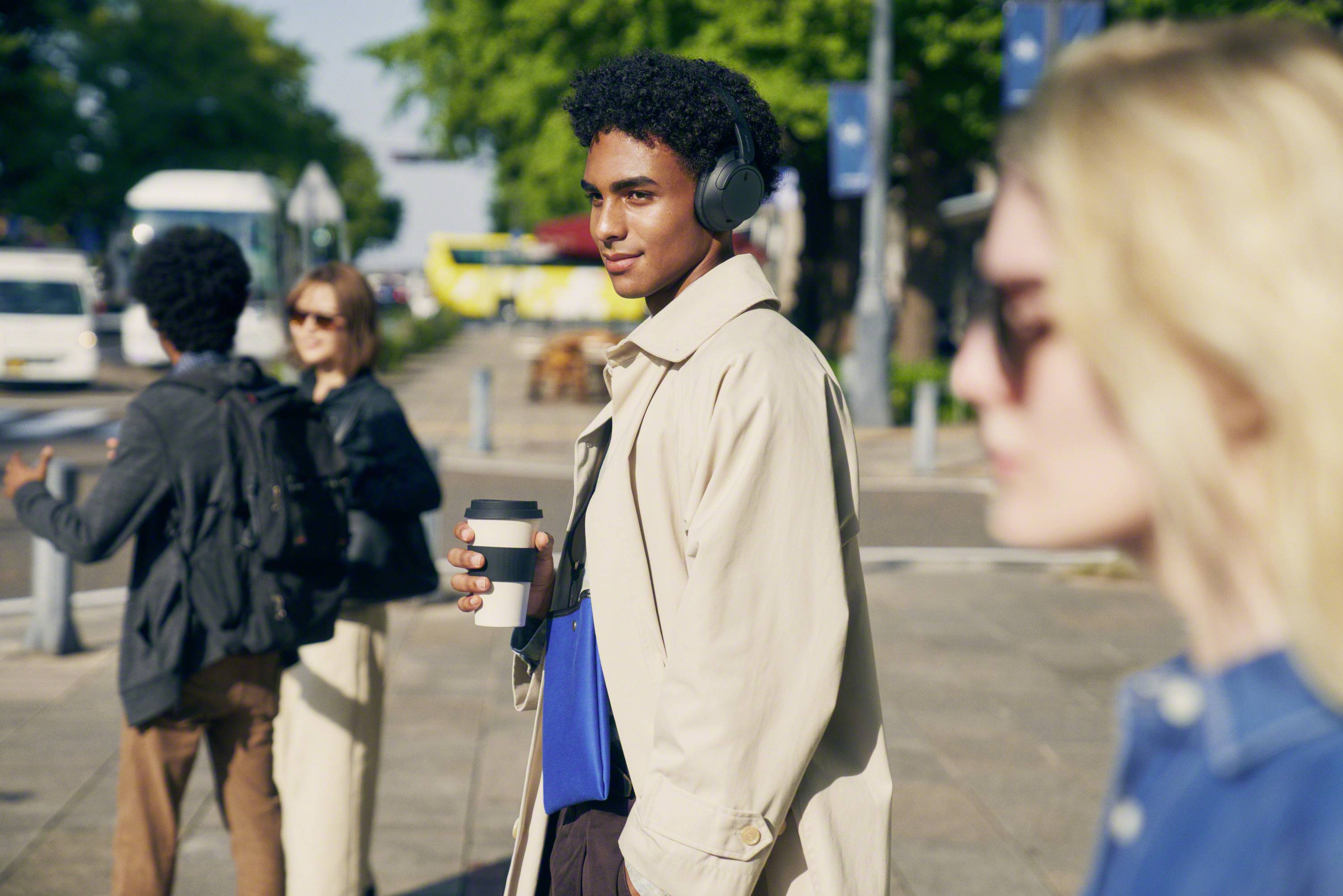 Une personne portant des écouteurs marche le long d'une rue animée, tenant un café et portant une pochette. D'autres personnes sont floues en arrière-plan.