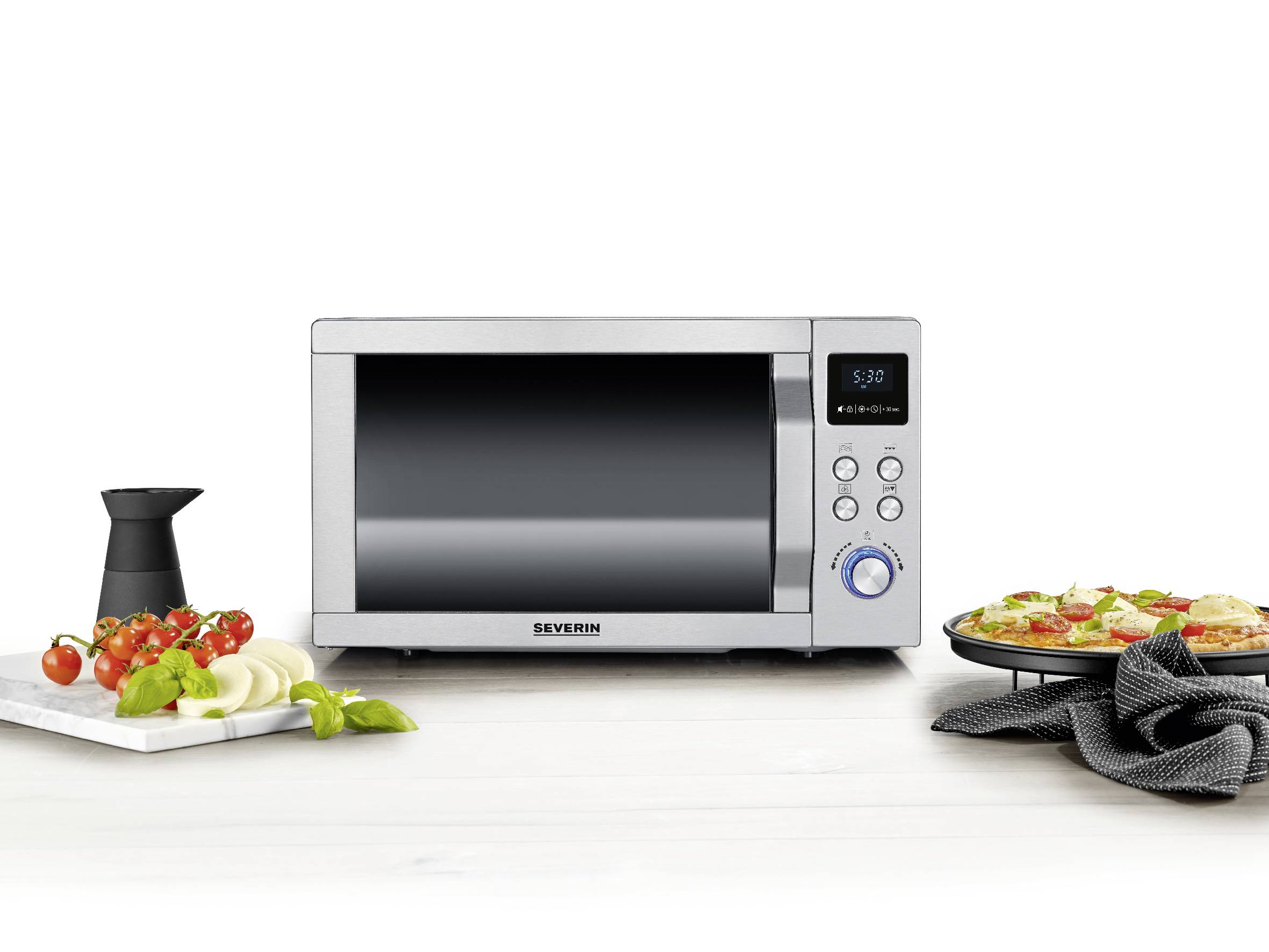 Four micro-ondes argenté sur une table de cuisine, entouré de tomates empilées, d'oignons, de basilic et d'une pizza dans une assiette.