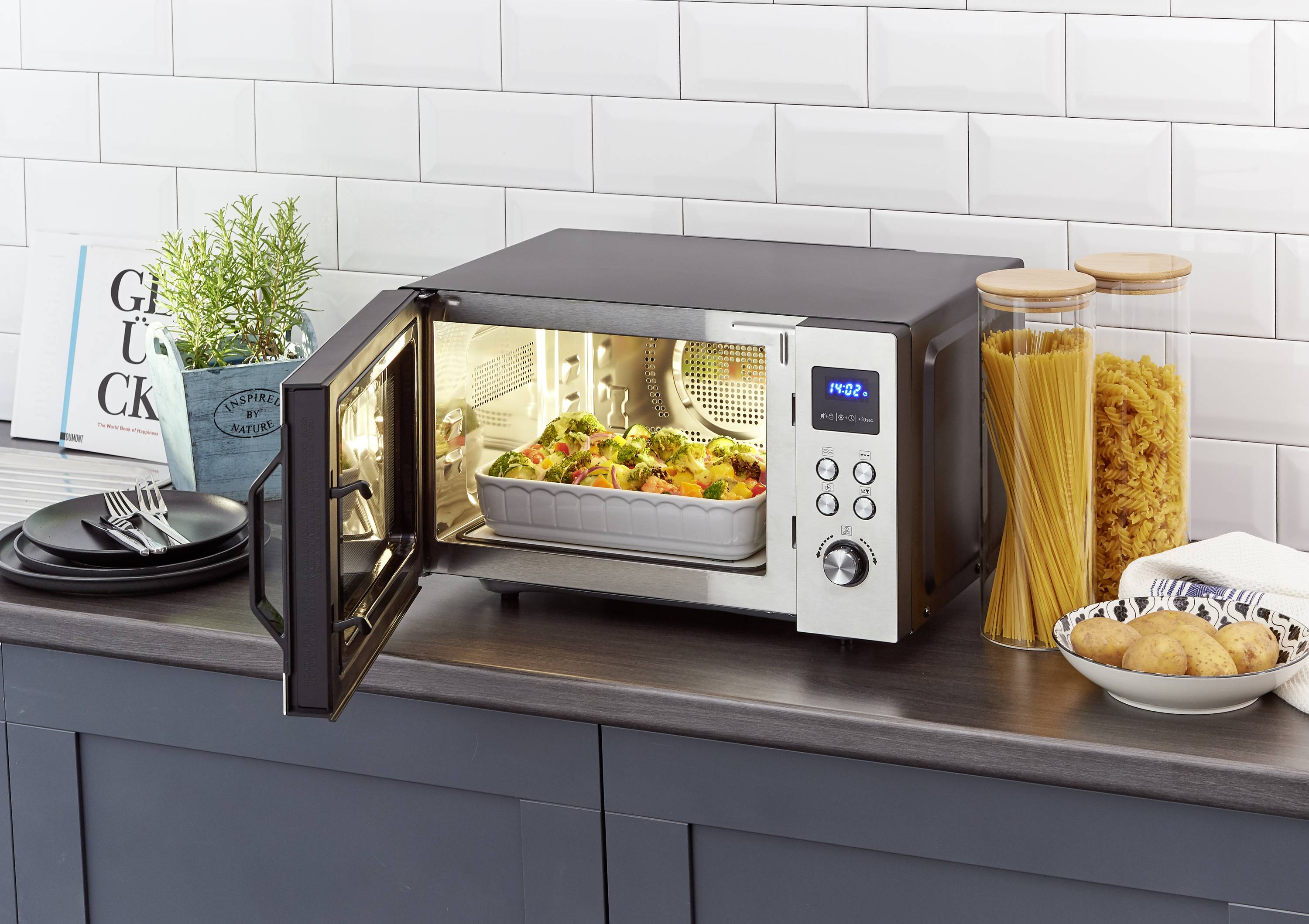 Une micro-ondes ouverte sur un plan de travail de cuisine, contenant un gratin. À côté, des récipients en verre remplis de pâtes et quelques assiettes sont disposés.