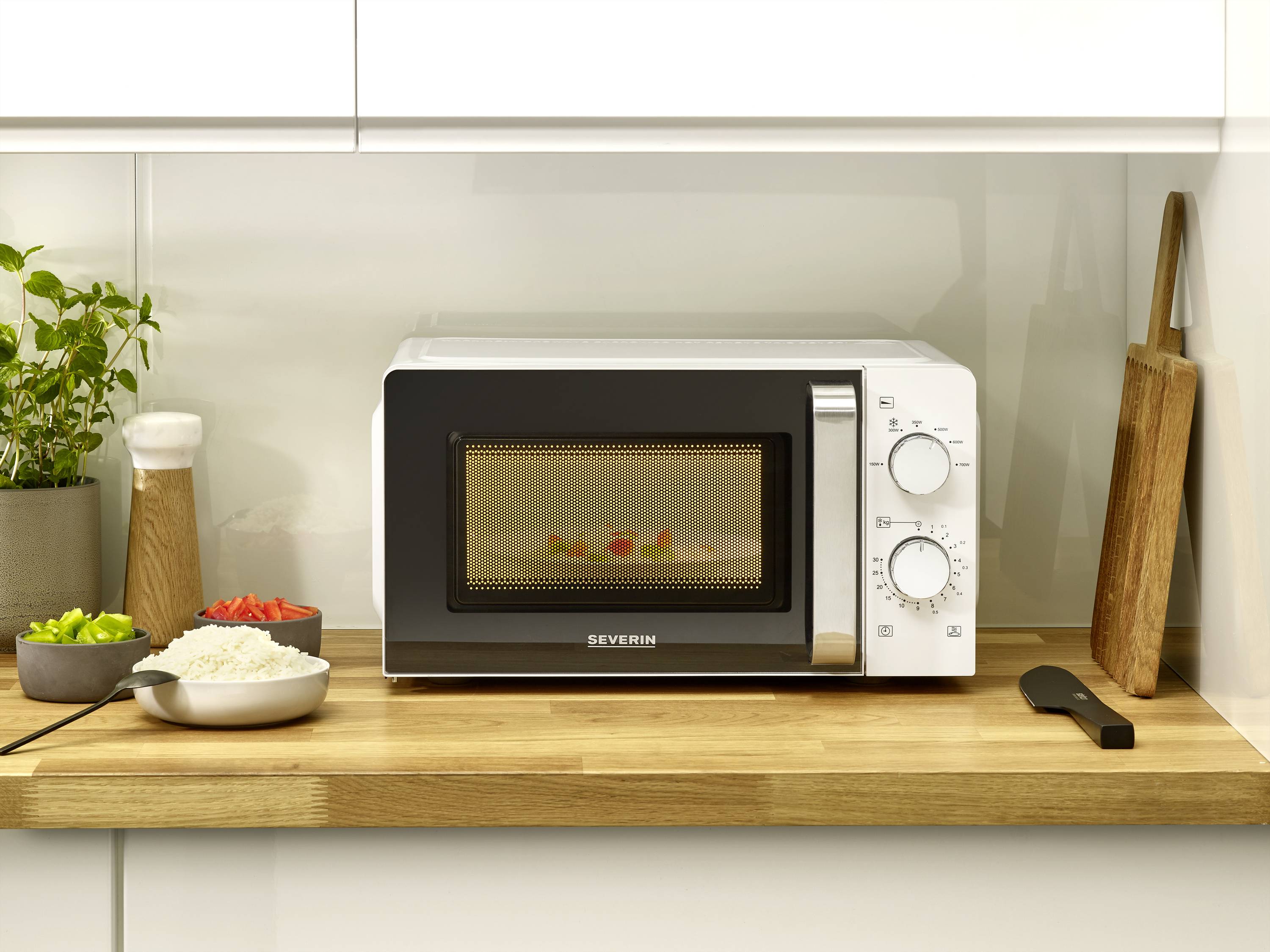 Four à micro-ondes sur un plan de travail en bois, à côté des épices et de la vaisselle ; à l'intérieur, des aliments sur une assiette. Cuisine avec armoire blanche.