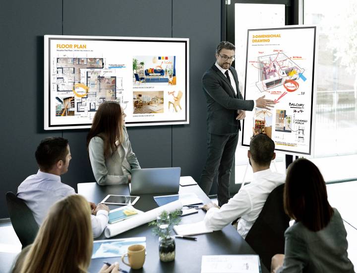 Un homme d'affaires présente des plans de construction et des dessins 3D sur des écrans à une équipe dans une salle de conférence.