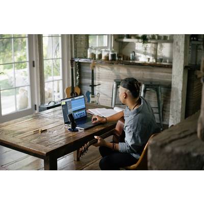 Un homme est assis à une table en bois avec un ordinateur portable et un microphone, jouant de la guitare. Une fenêtre en arrière-plan donne sur un jardin ensoleillé. Un espace de travail créatif.