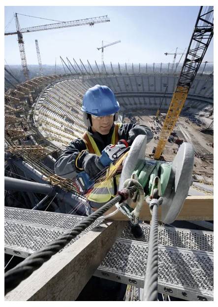 Un ouvrier du bâtiment portant un équipement de sécurité attache des cordes sur une poutre métallique sur un grand chantier de construction de stade, entouré de grues et d'équipements.