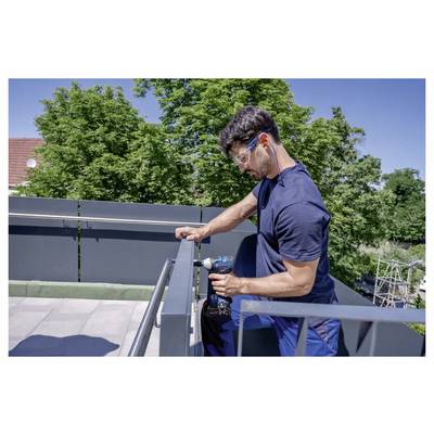 Une personne sur un balcon de toit utilisant une perceuse électrique pour fixer une rambarde métallique, avec des arbres en arrière-plan.