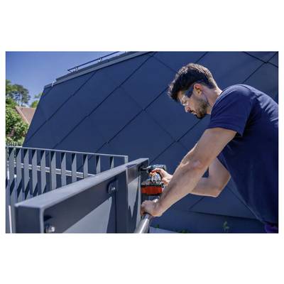 Une personne portant des lunettes de sécurité et une protection auditive utilise une perceuse pour travailler sur une rambarde métallique à l'extérieur, avec un bâtiment moderne en arrière-plan.