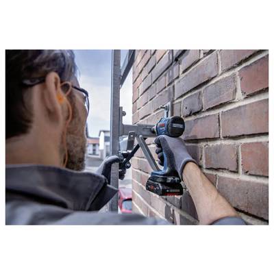 Une personne équipée d'un équipement de sécurité utilise une perceuse sans fil pour fixer un élément métallique sur un mur de briques, concentrée sur des travaux de construction ou de réparation.
