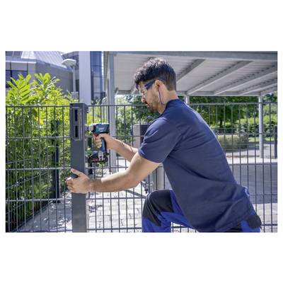 Un homme portant des lunettes de protection utilise une perceuse électrique pour fixer un portail métallique à l'extérieur par une journée ensoleillée.