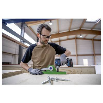 Un homme portant des lunettes de sécurité et des gants utilise une perceuse électrique sur une pièce métallique verte dans un atelier de menuiserie avec des poutres et des vis visibles.