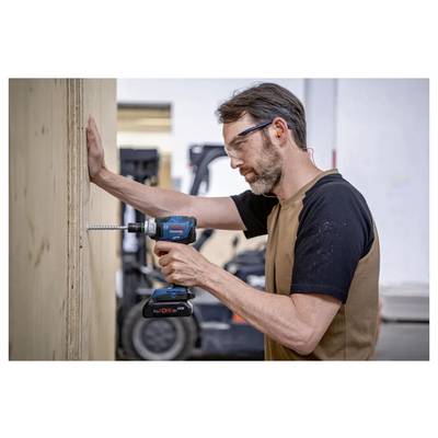 Une personne dans un atelier utilise une perceuse sans fil bleue pour visser une longue vis dans une planche en bois, avec un chariot élévateur visible en arrière-plan.