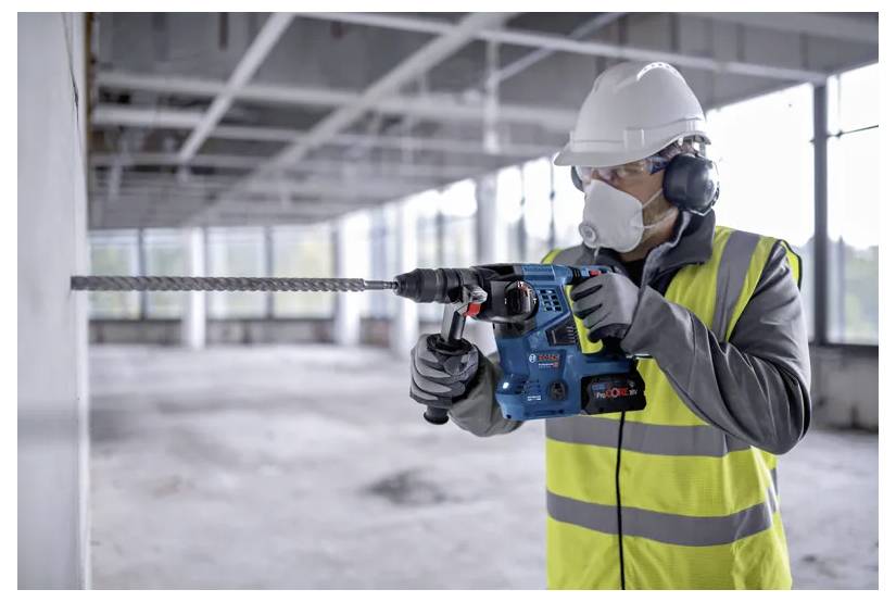 Un travailleur portant un équipement de protection utilise une perceuse électrique sur un mur en béton dans un grand bâtiment inachevé.