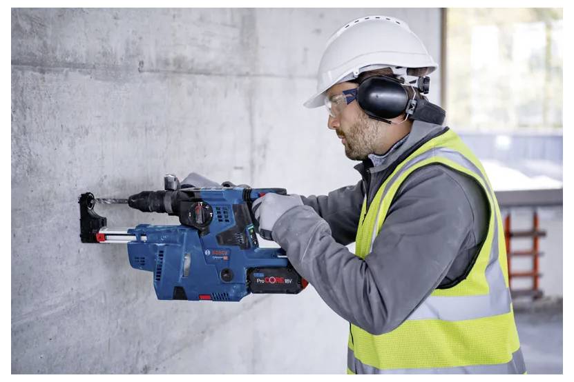 Un travailleur, portant un équipement de sécurité, utilise un marteau perforateur rotatif bleu sur un mur en béton à l'intérieur d'un chantier de construction.