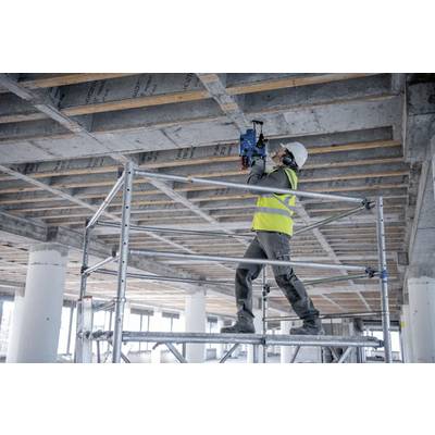 Un ouvrier du bâtiment portant un casque de chantier et un gilet de sécurité utilise un outil électrique sur un plafond en béton, debout sur un échafaudage dans une structure en cours de construction.
