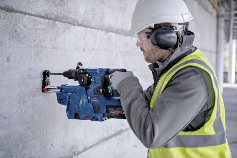 Un ouvrier du bâtiment portant un équipement de sécurité utilise une perceuse électrique bleue pour percer un mur en béton.