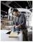 Un homme portant des lunettes de sécurité utilise une scie électrique, coupant une planche en bois dans un environnement d'atelier industriel.