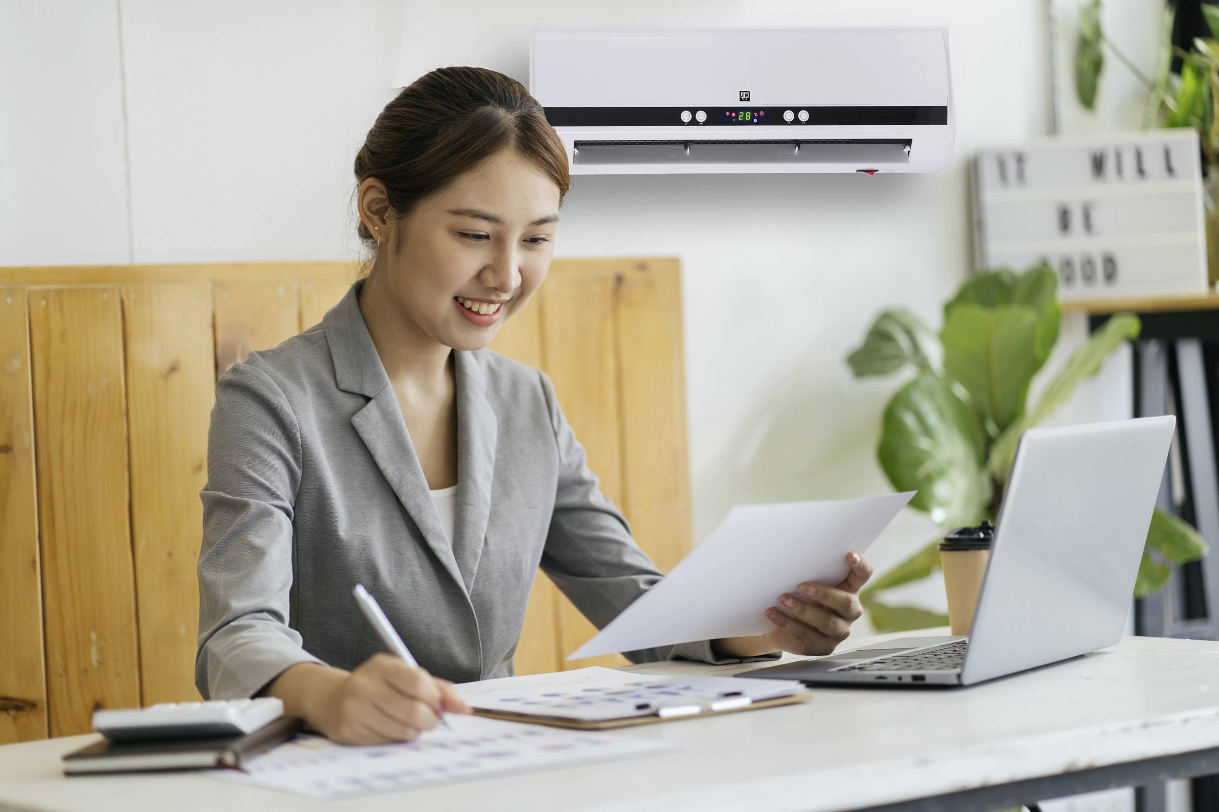 Une femme en tenue de bureau travaille sur un ordinateur portable et examine un document. Un climatiseur est accroché au mur en arrière-plan.