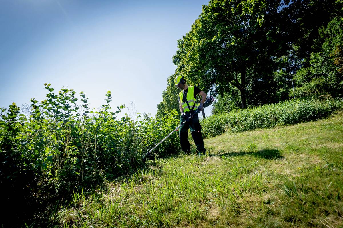 Une personne portant un gilet de sécurité coupe l'herbe avec un coupe-bordures sur une prairie. Des arbres et un ciel bleu sont visibles en arrière-plan.