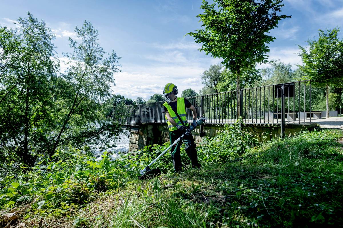 Une personne en tenue de travail coupe de l'herbe et des mauvaises herbes avec une débroussailleuse près d'une rivière, avec un pont en arrière-plan.