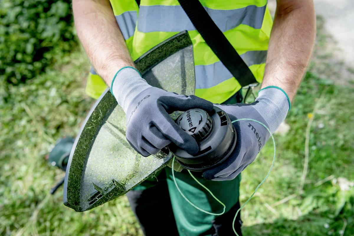 Une personne porte des gants et tient un taille-haie, prête à l'utiliser dans un jardin. De l'herbe et des plantes vertes sont visibles en arrière-plan.
