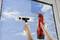 Une personne nettoie une fenêtre avec un produit de nettoyage en spray et un raclette à vitres. Le ciel se reflète dans la vitre, les nuages sont visibles.