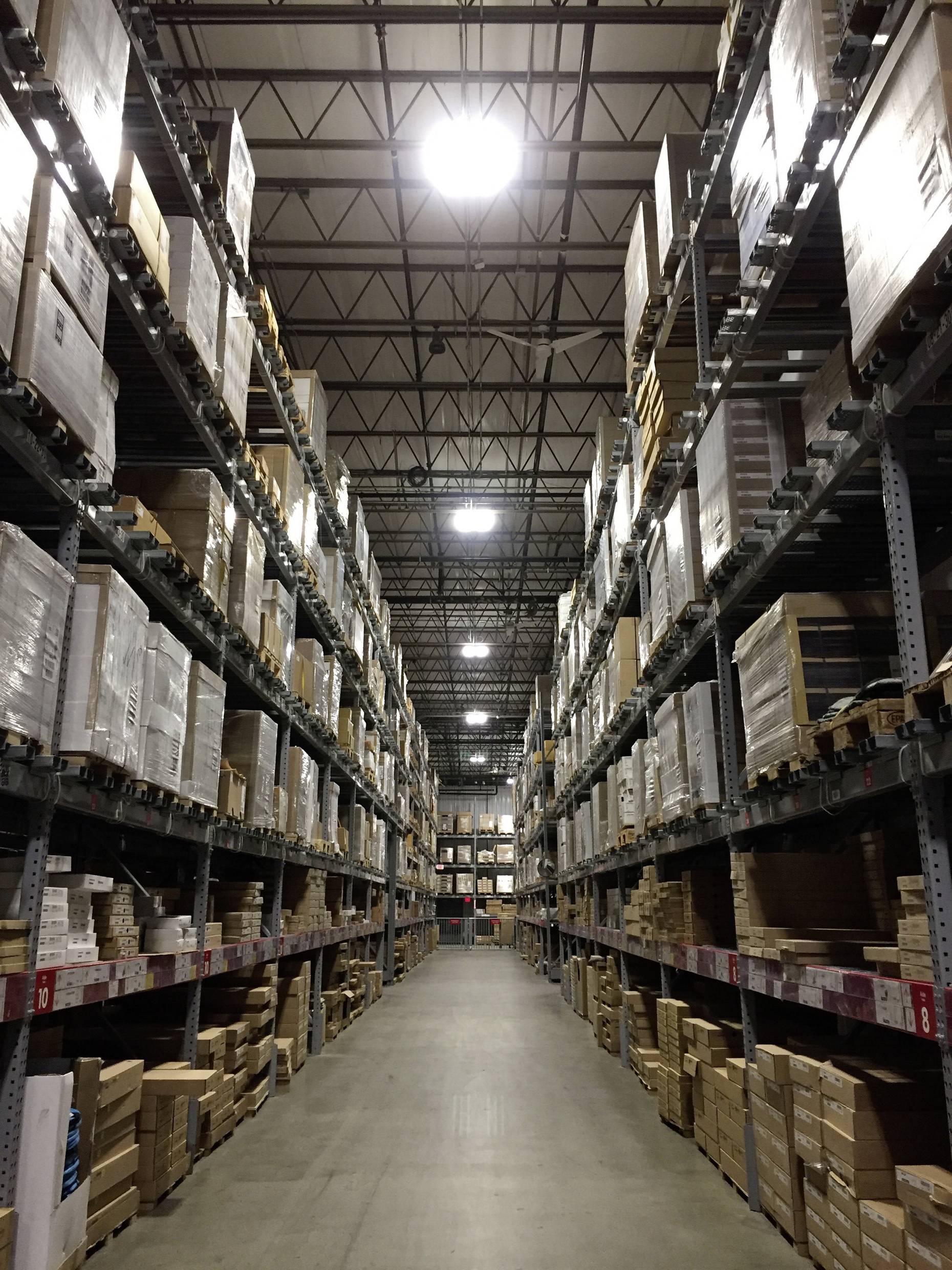 Un grand entrepôt avec des étagères hautes remplies de cartons et de palettes empilés. Un long couloir traverse le centre du hall.