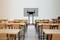 Salle de classe avec des tables et des chaises en bois vides. Un grand écran à l'avant. Des murs sont visibles à gauche et à droite, ainsi qu'un bureau d'enseignant.