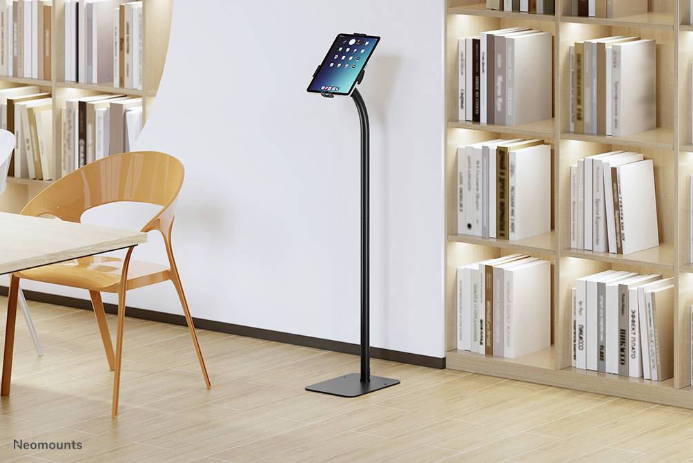 'Un support de tablette moderne avec une tablette, entouré de étagères à livres et d'une table avec une chaise orange, dans une pièce lumineuse.'