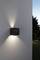 Applique murale noire en forme de cube sur un mur blanc, projetant de la lumière vers le haut et vers le bas, jardin en arrière-plan au crépuscule.