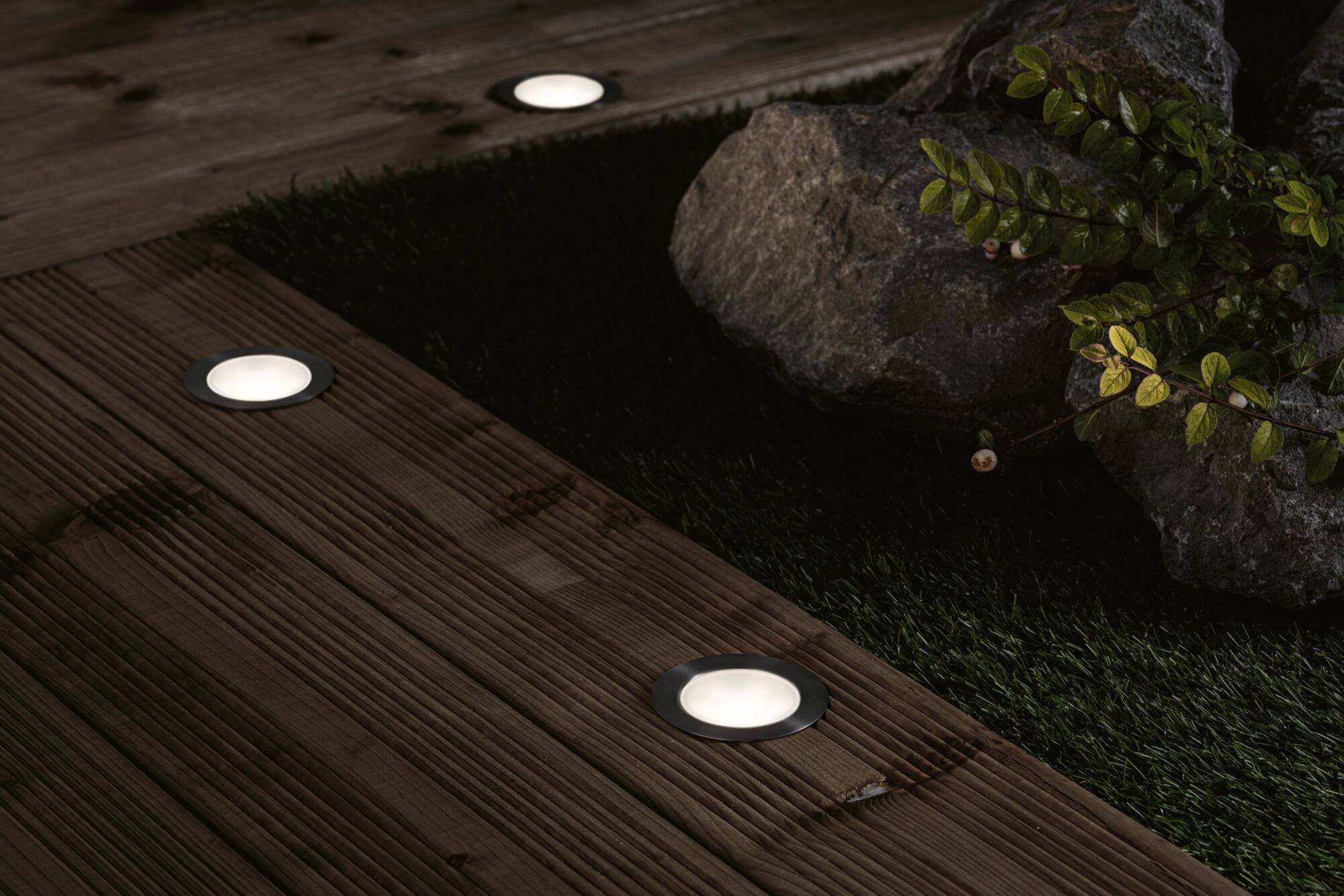 Terrasse en bois avec des luminaires encastrés au sol, entourée de pierres et de plantes.