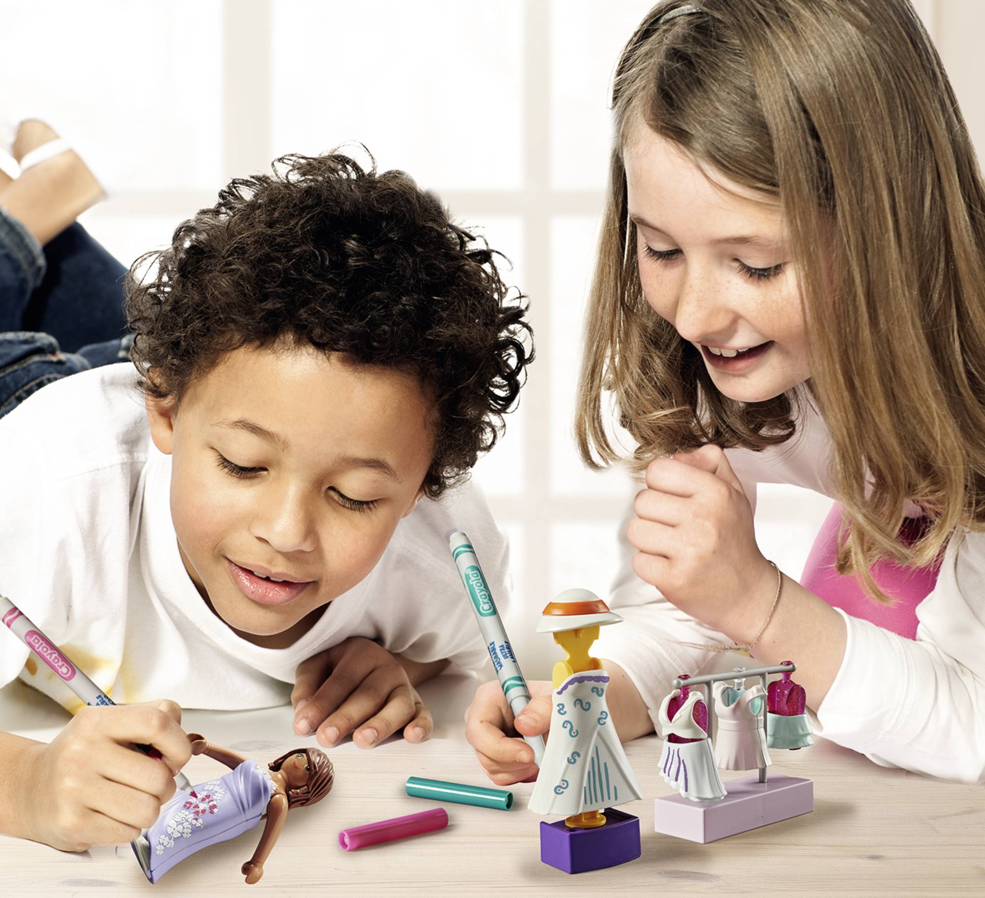 Deux enfants sont allongés au sol et dessinent des figures avec des crayons. Ils semblent concentrés et créatifs.