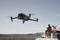 Un drone survole un paysage désertique, tandis qu'un homme assis dans un tout-terrain avec un chien contrôle la télécommande.
