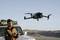 Un homme pilote un drone à l'aide d'une télécommande devant un véhicule tout-terrain dans un paysage désertique. Le drone est suspendu dans les airs.
