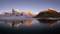 Une chaîne de montagnes aux sommets enneigés se reflète dans un lac calme au premier plan, illuminée par le coucher de soleil.