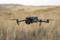 Un drone survole un vaste paysage aride couvert d'herbes hautes. En arrière-plan, des collines floues baignent dans la lumière du soleil.
