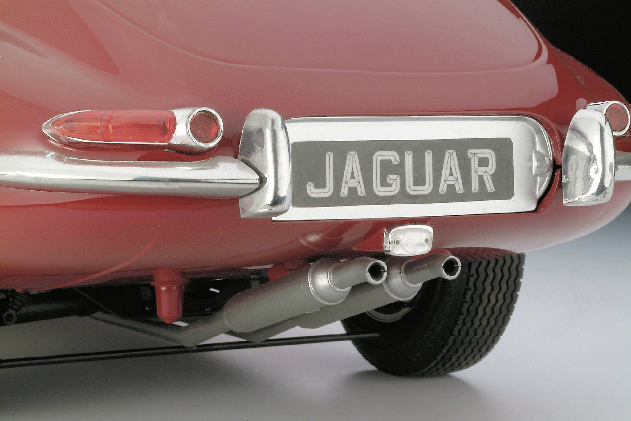 Arrière d'une voiture rouge avec un emblème Jaguar sur la plaque d'immatriculation, doubles tuyaux d'échappement et feux arrière ronds. Design rétro.