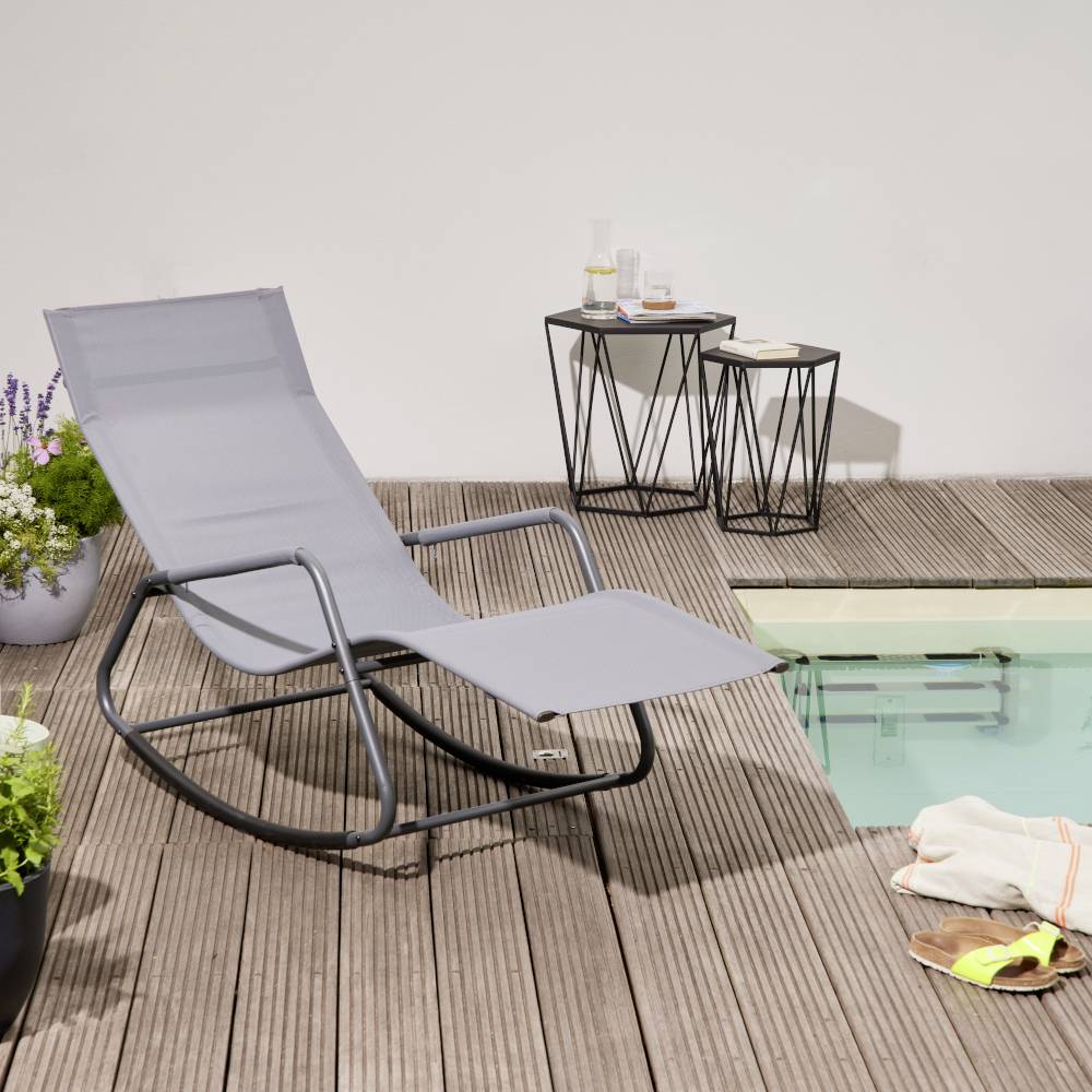 Un fauteuil à bascule gris est placé sur une terrasse en bois à côté d'une petite table d'appoint avec des boissons, donnant sur une piscine.