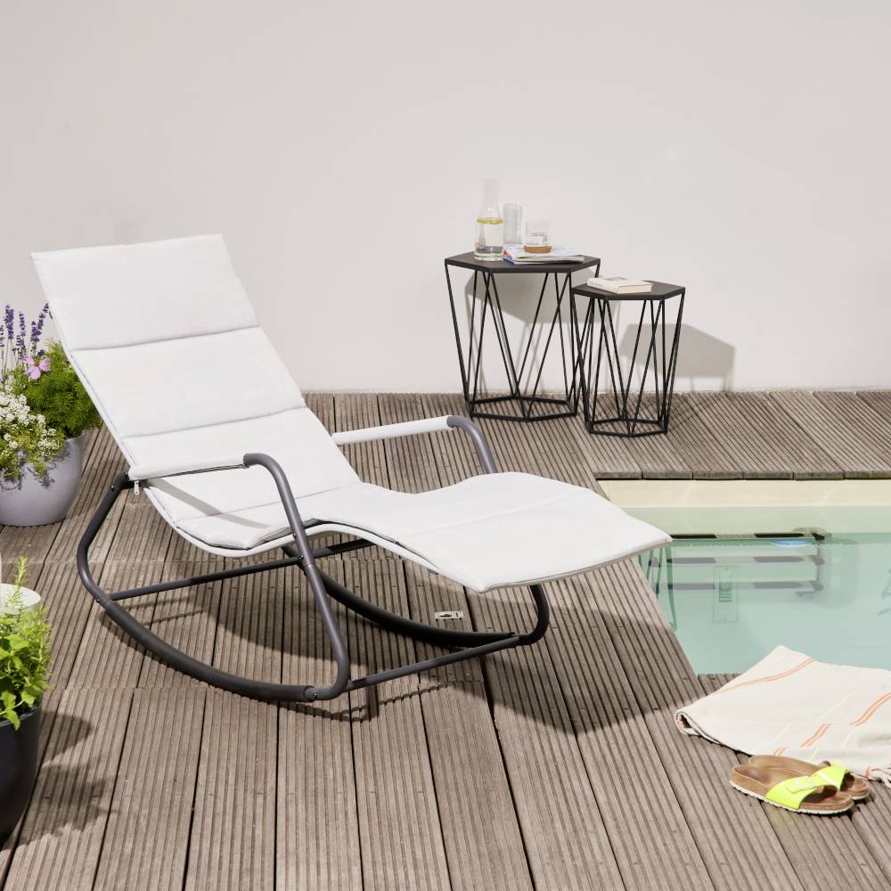 Chaise longue et tables d'appoint sur une terrasse en bois à côté d'une piscine. Une serviette est posée sur la terrasse, des fleurs en arrière-plan.