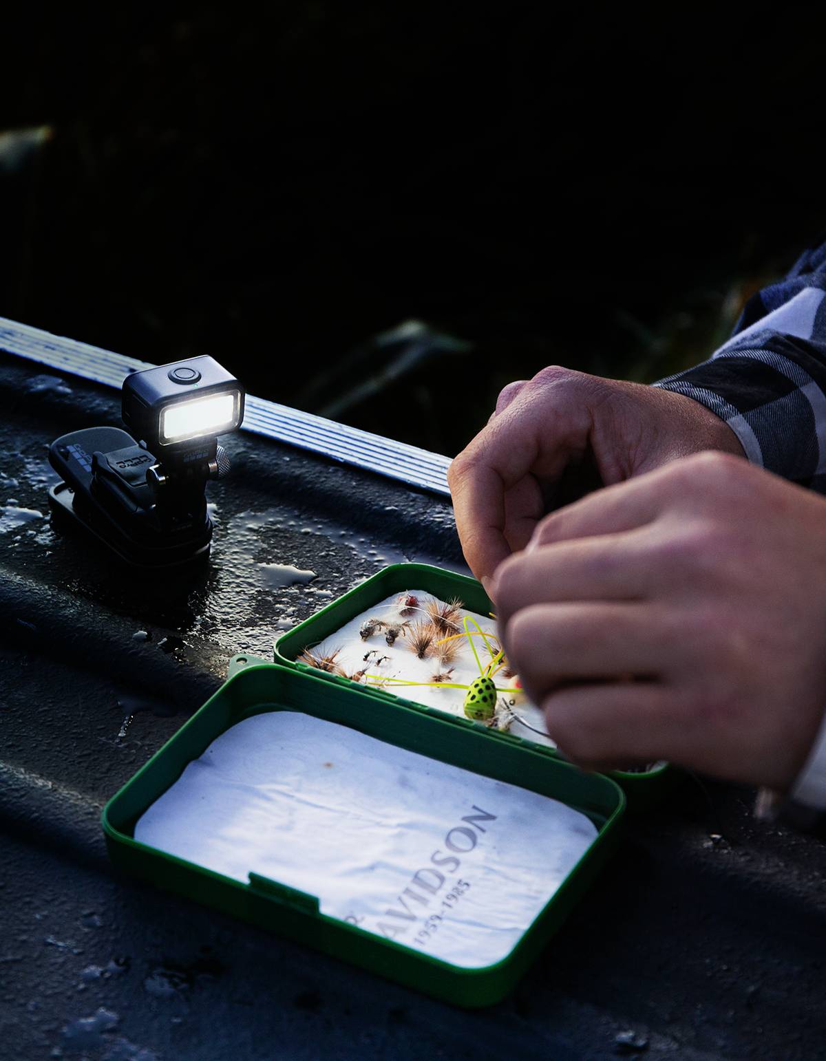 Une personne prépare des appâts de pêche dans une boîte verte. Une petite lampe de poche brille à côté, il fait noir dehors.