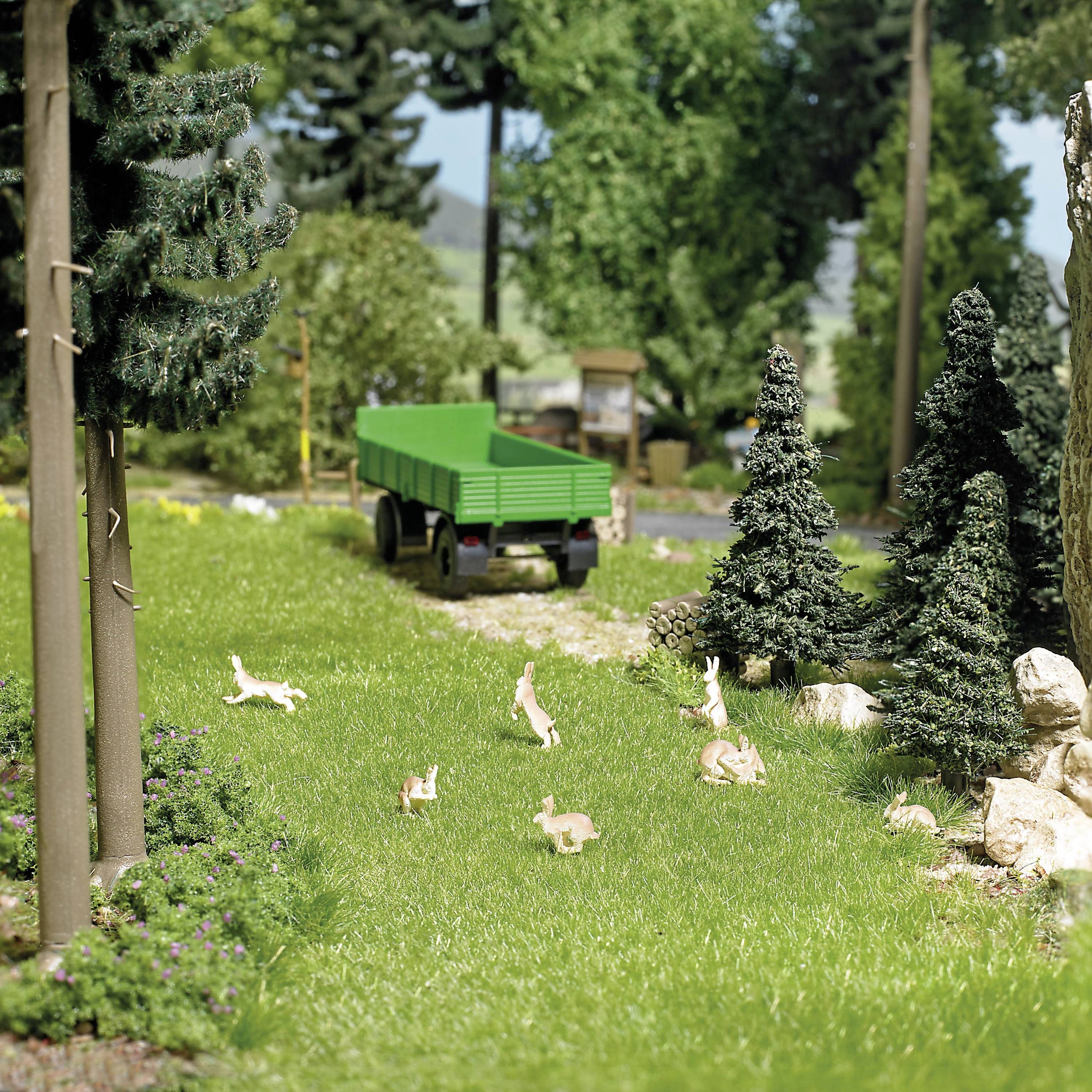 Paysage miniature avec une remorque de tracteur verte, un chemin couvert d'herbe et des sapins, au milieu de figurines de lapins sur une prairie.