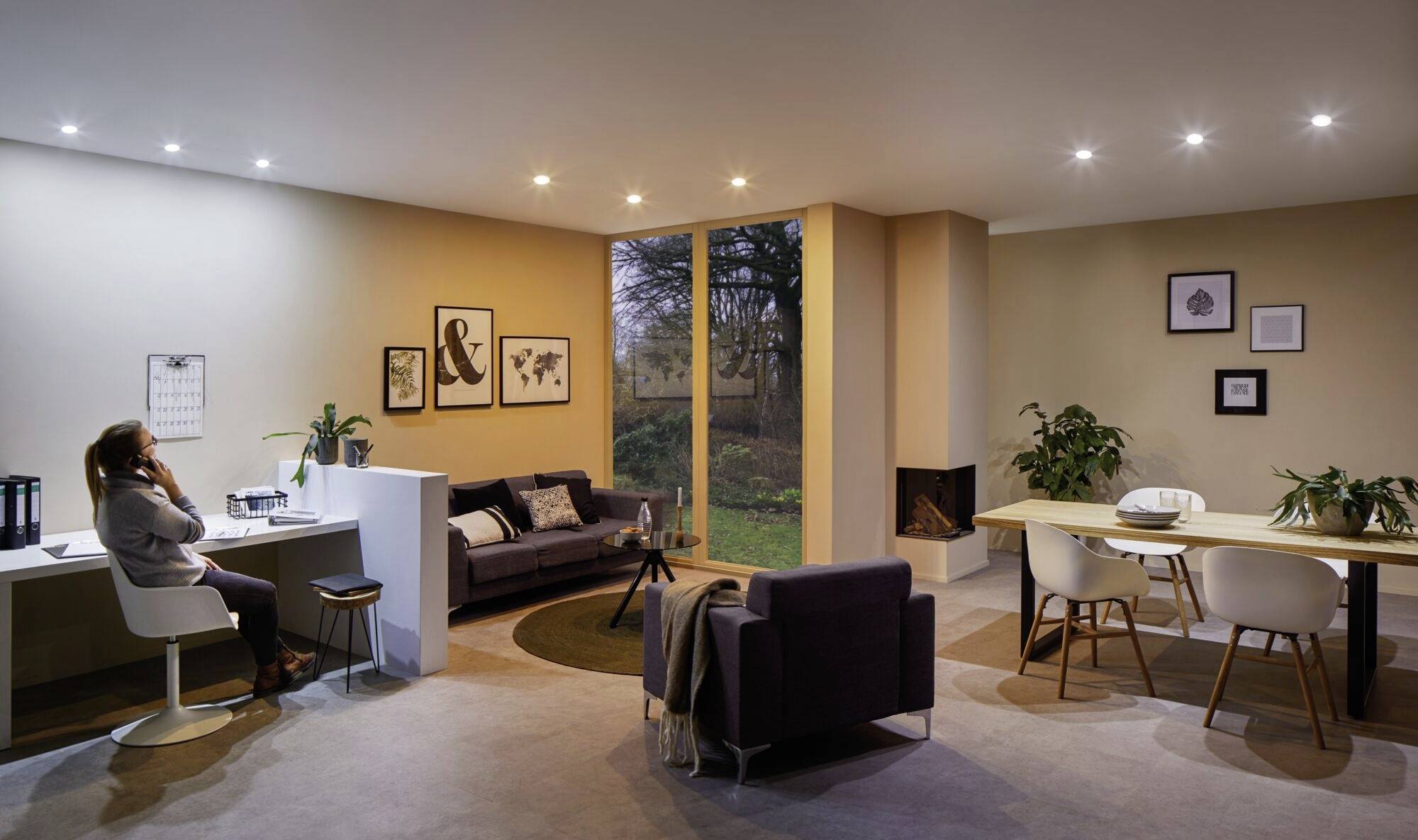 Une personne téléphone au bureau dans un salon moderne, avec deux canapés à proximité, une table à manger et des chaises, des plantes et des tableaux accrochés aux murs.
