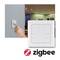Une main actionne un interrupteur mural. À côté se trouve un interrupteur domotique intelligent avec le logo Zigbee. En arrière-plan, une pièce éclairée.