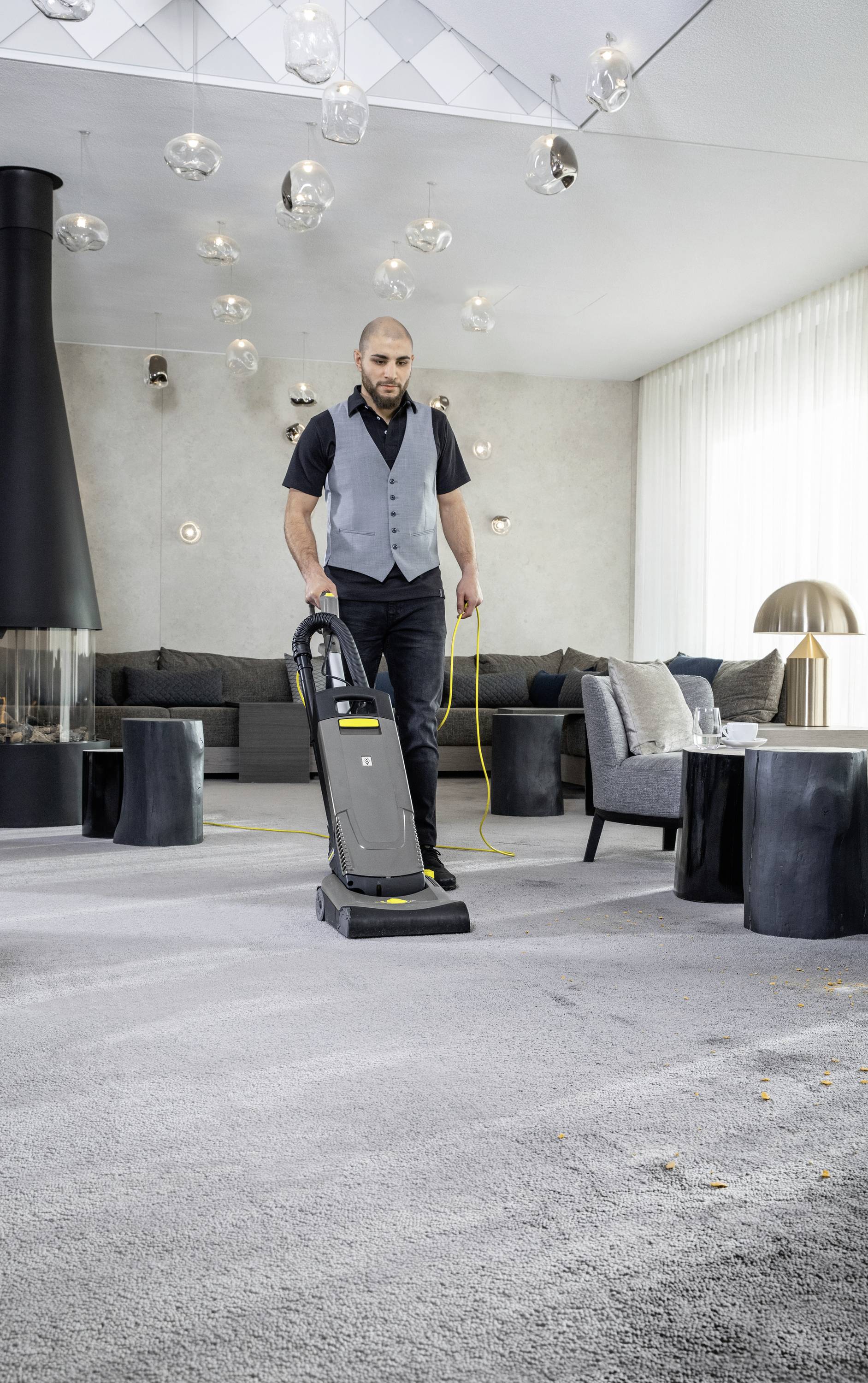 Une personne aspire un tapis gris avec un aspirateur dans un salon moderne doté d'une cheminée et de canapés.